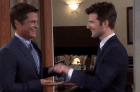 Two men in suits engage in a light-hearted handshake, smiling and pointing at each other as they exchange a friendly gesture in an office setting