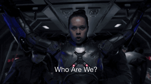 A woman in a futuristic, armored suit is inside a spaceship with arms raised, asking with determination, "Who are we?" The background shows a dimly lit cockpit, adding intensity to the scene.