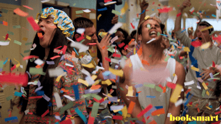 A group of excited people are celebrating with confetti flying in the air, cheering and shouting in a hallway. One person is wearing a shower cap, and the scene is vibrant and lively. The text "booksmart" appears in the bottom right corner