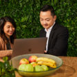 Two people working together on a laptop at a table with a bowl of fruit, set against a green leafy background