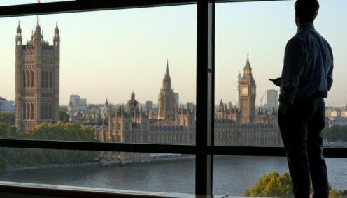 A person stands by a large window overlooking the iconic British Houses of Parliament and Big Ben in the distance, silhouetted against the early morning or late afternoon light, creating a contemplative mood