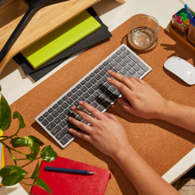 Person typing on a keyboard at a tidy desk with a mouse, notebooks, and a plant nearby