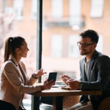 Two colleagues sitting at a table in a cafe, engaging in conversation and smiling, with one holding a smartphone
