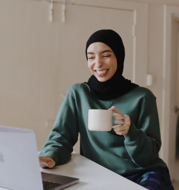 Smiling person wearing a black hijab and green sweater, holding a white mug and looking down, enjoying a moment of laughter.
