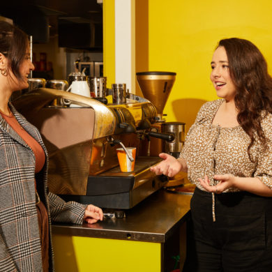 The image shows two people engaged in a conversation in front of a coffee machine, set against a yellow background.