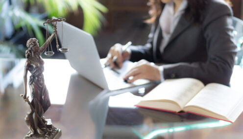 A close-up of a justice statue on a glass desk with a woman in a business suit working on a laptop in the background, next to an open book