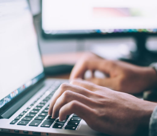 Close-up of hands typing on a laptop keyboard, with a monitor visible in the background