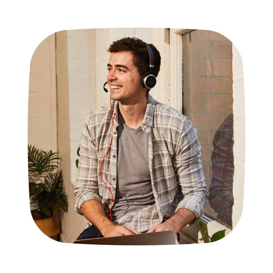 Person wearing a headset and smiling, sitting near a window with a laptop, in a casual setting with indoor plants