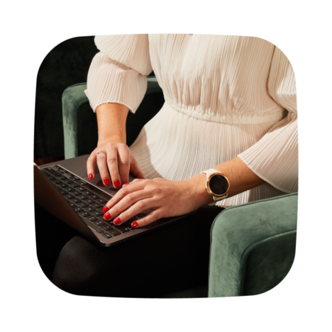 Person typing on a laptop while seated in a green chair, wearing a white blouse and a smartwatch
