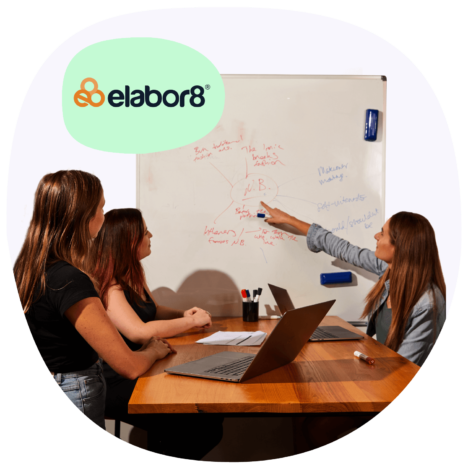A photo of three women sitting around a table, engaged in a discussion. One woman is pointing to a whiteboard with notes and diagrams