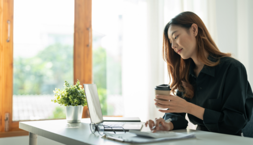 A woman sitting at a desk with a laptop, holding a coffee cup, looking focused while working in a well-lit room