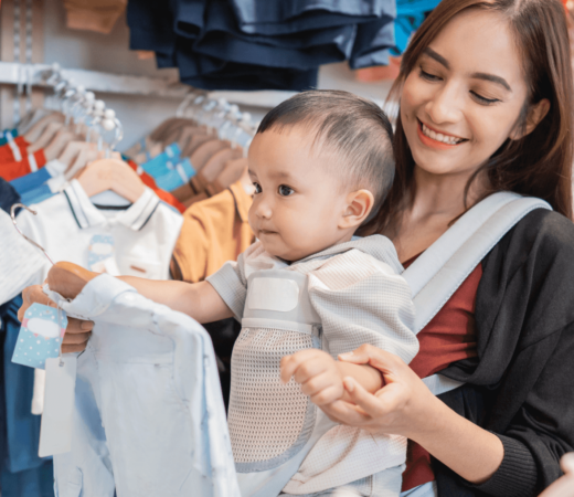 Mom shopping in a store while holding up a piece of baby's clothing in front of her kid in a carrier
