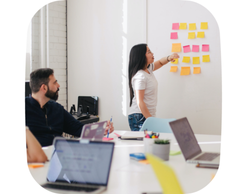 A photo of a woman standing and pointing to sticky notes on a wall during a meeting, while others sit at a table with laptops