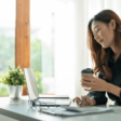 A woman sits at a desk in a bright office, holding a coffee cup while looking at a laptop