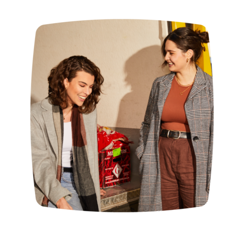 A photo of two women standing and smiling, engaged in a friendly conversation, dressed in casual yet stylish outfits, set against an urban backdrop