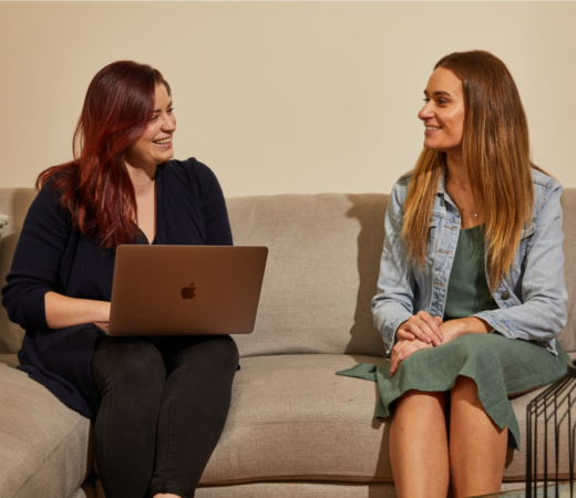 Two women sitting on a couch, smiling and talking, with one holding a laptop