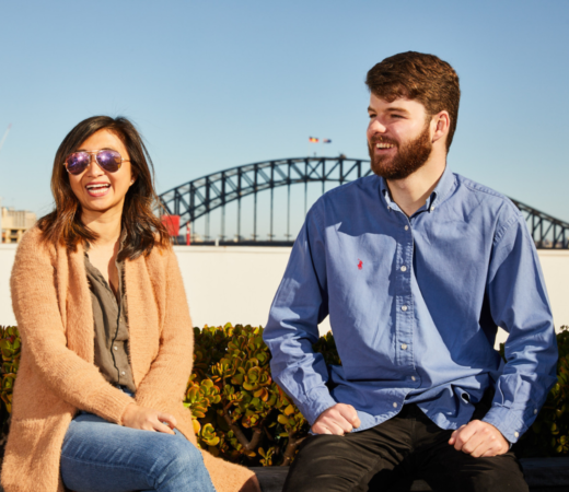 A cheerful outdoor photo of two people, a woman and a man, sitting in front of the iconic Sydney Harbour Bridge