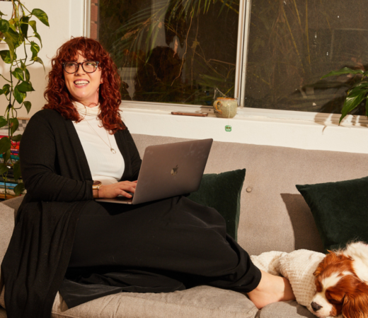 A woman with curly red hair and glasses sits comfortably on a couch, working on her laptop